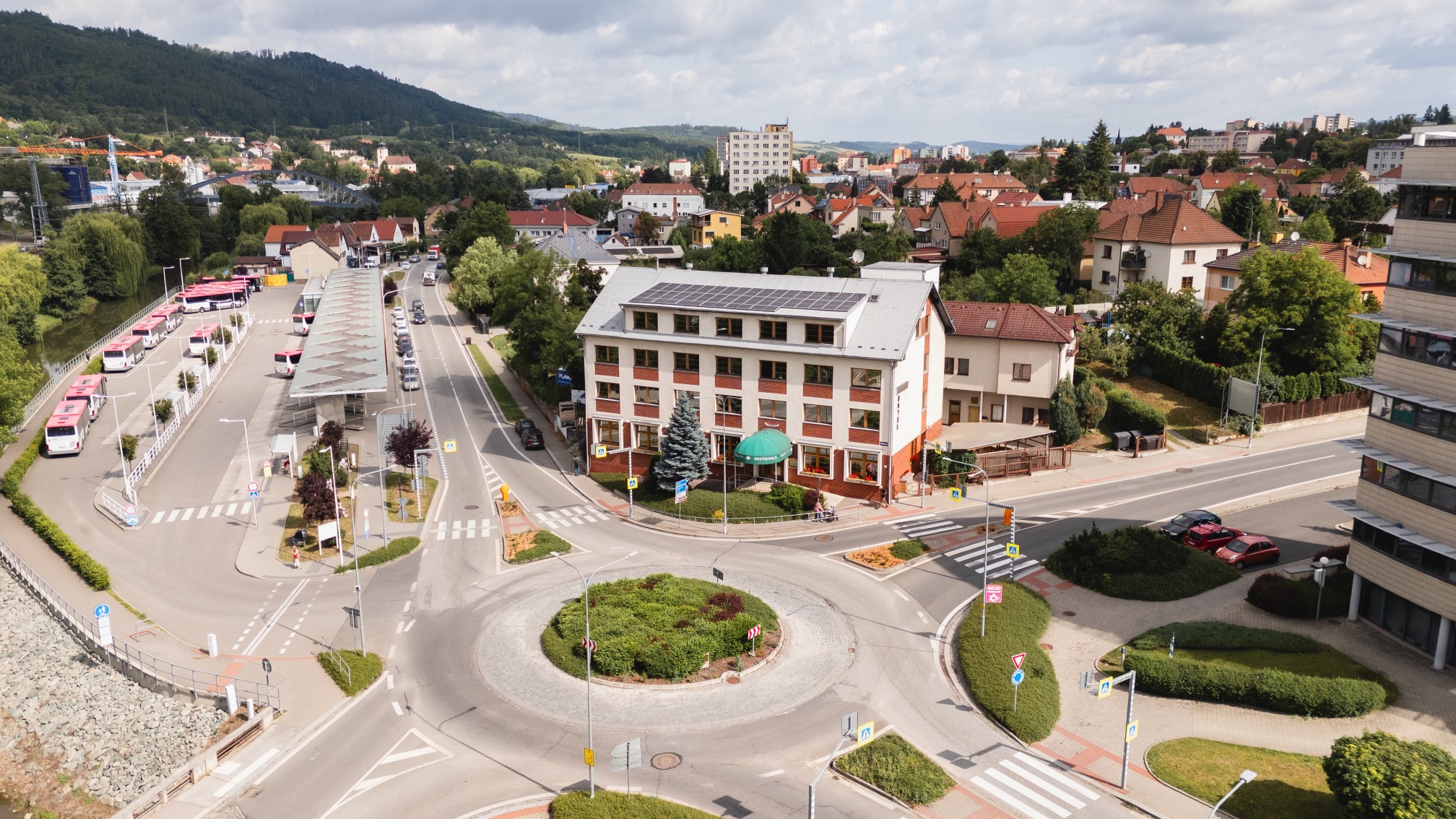 Aerial view of a roundabout surrounded by buildings and greenery, with a bus terminal featuring several red and white buses on the left and residential houses in the background.