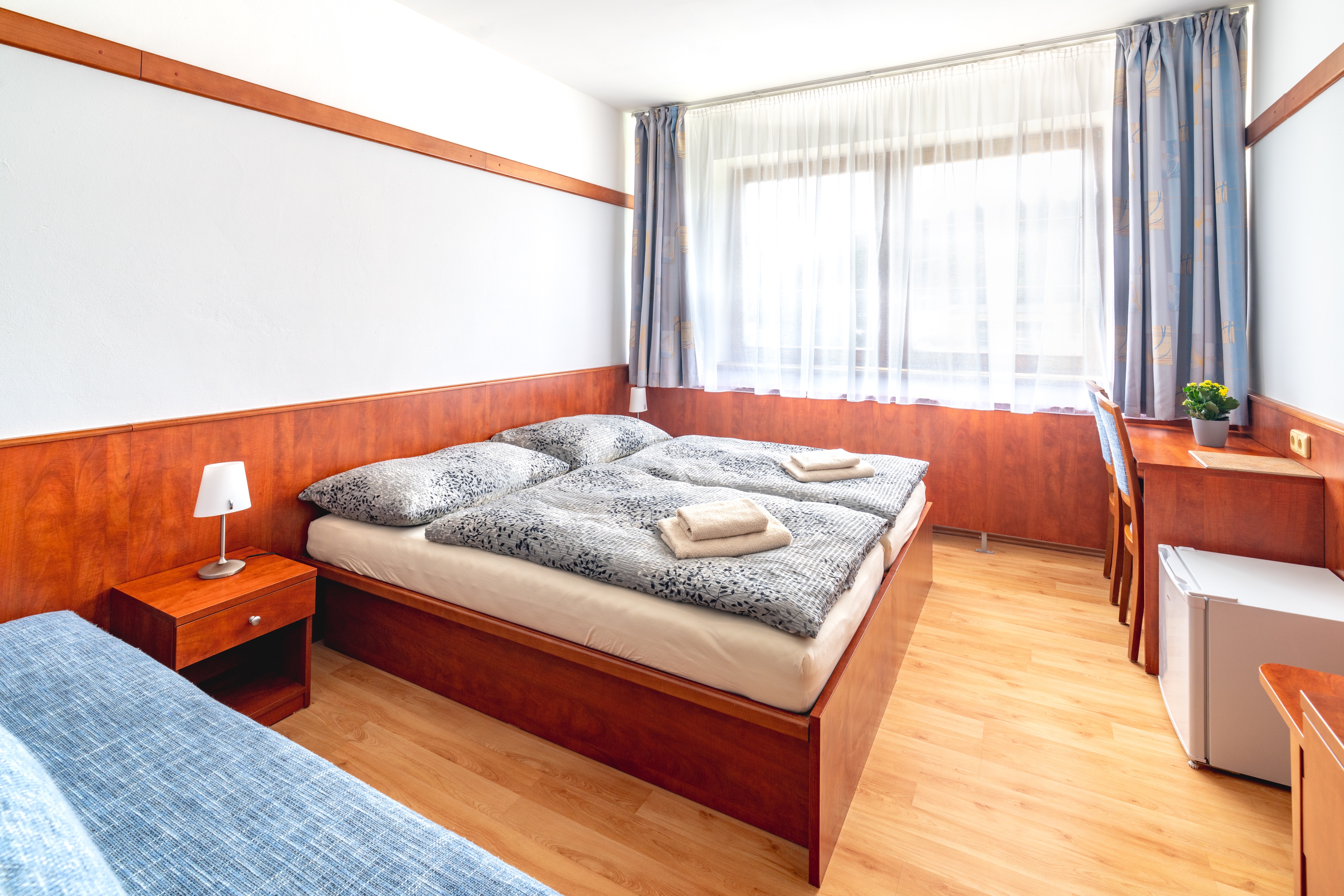 Bright hotel room with a wooden double bed covered with patterned gray bedding and folded towels, a bedside table with a lamp, a desk with a chair and a small refrigerator.