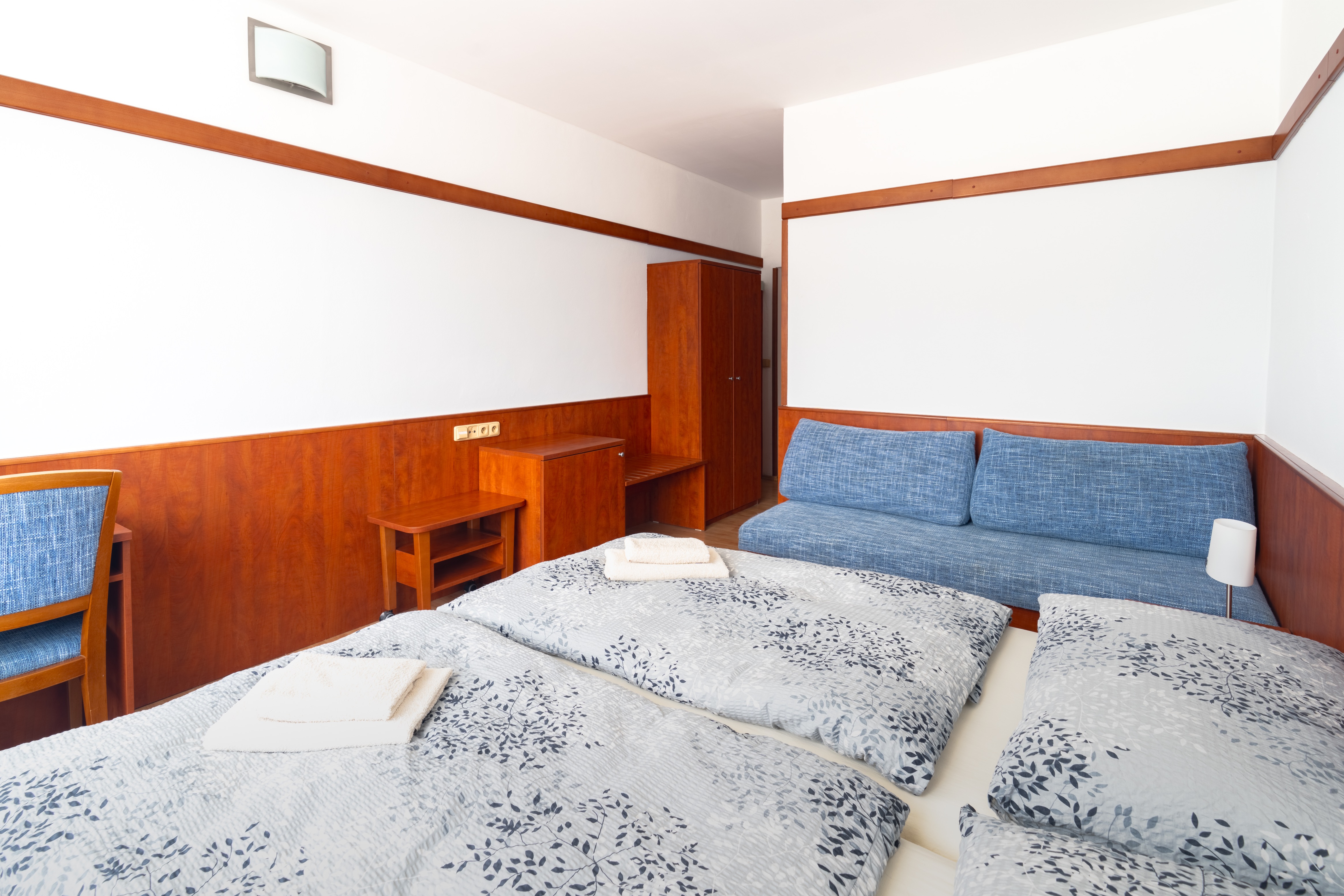 Bright hotel room with twin beds featuring patterned gray bedding, blue cushioned sofa, and wooden furniture.