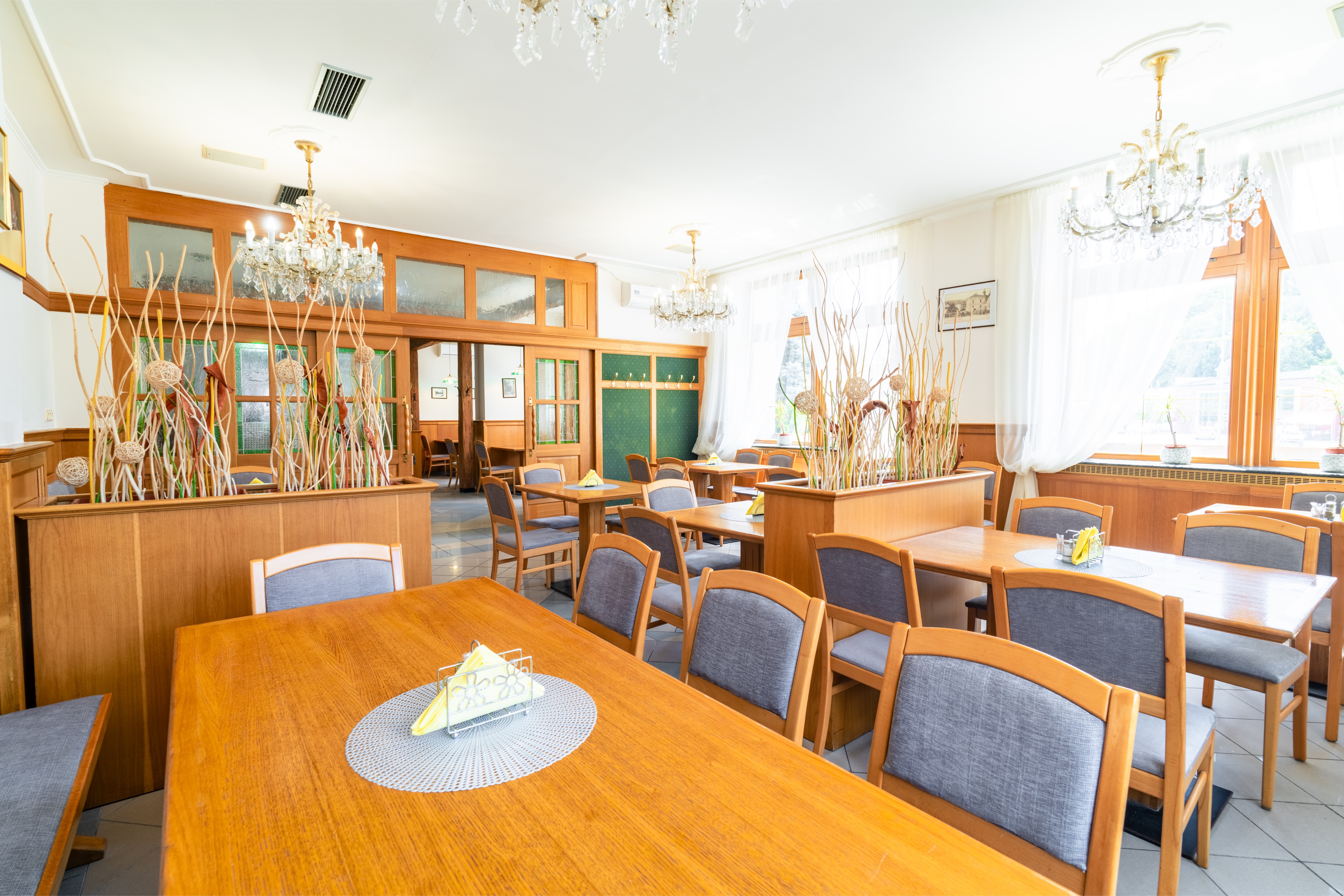 Bright dining room with wooden tables, gray cushioned chairs, white curtains, and decorative chandeliers.