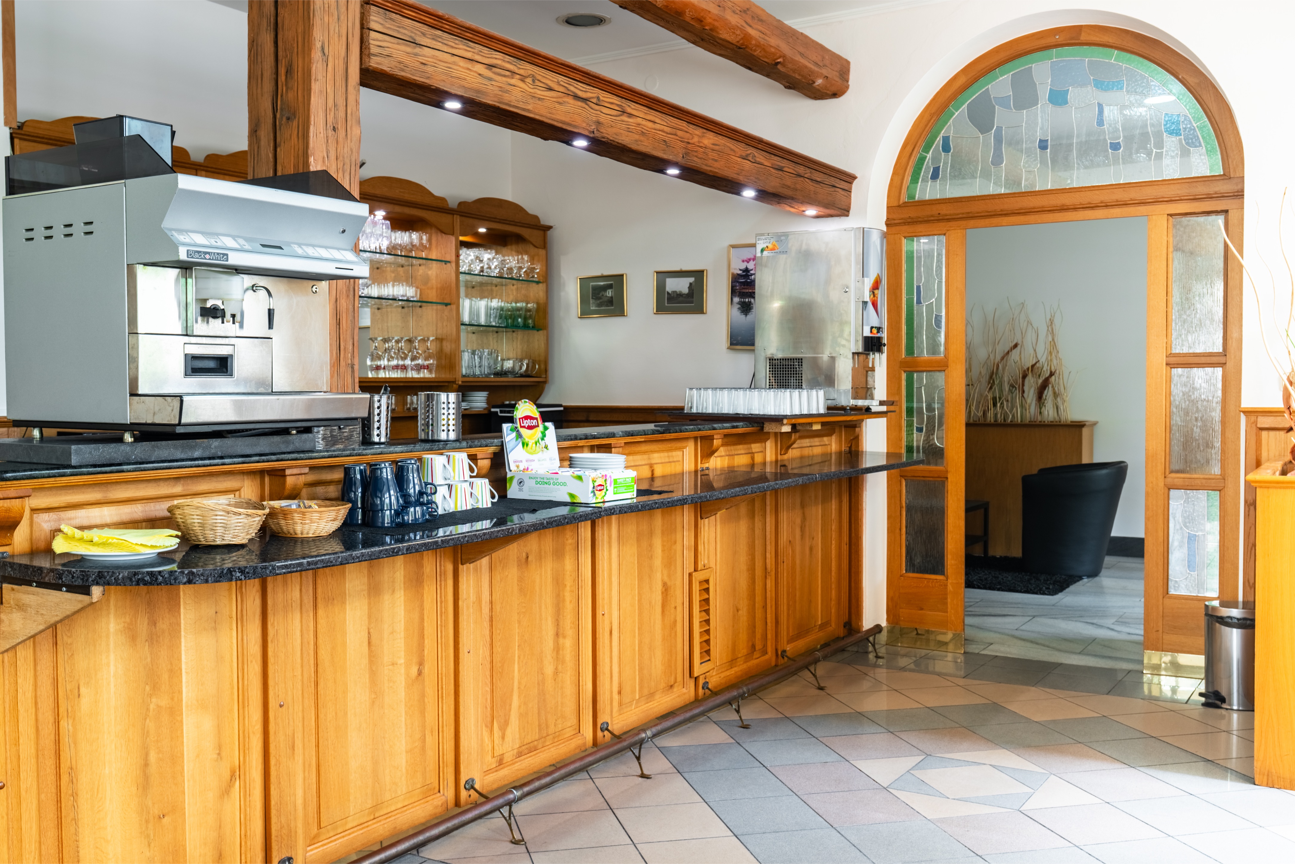 Wood-paneled coffee and tea bar with espresso machine, cups, glassware, and a beverage dispenser next to an arched doorway with stained glass.