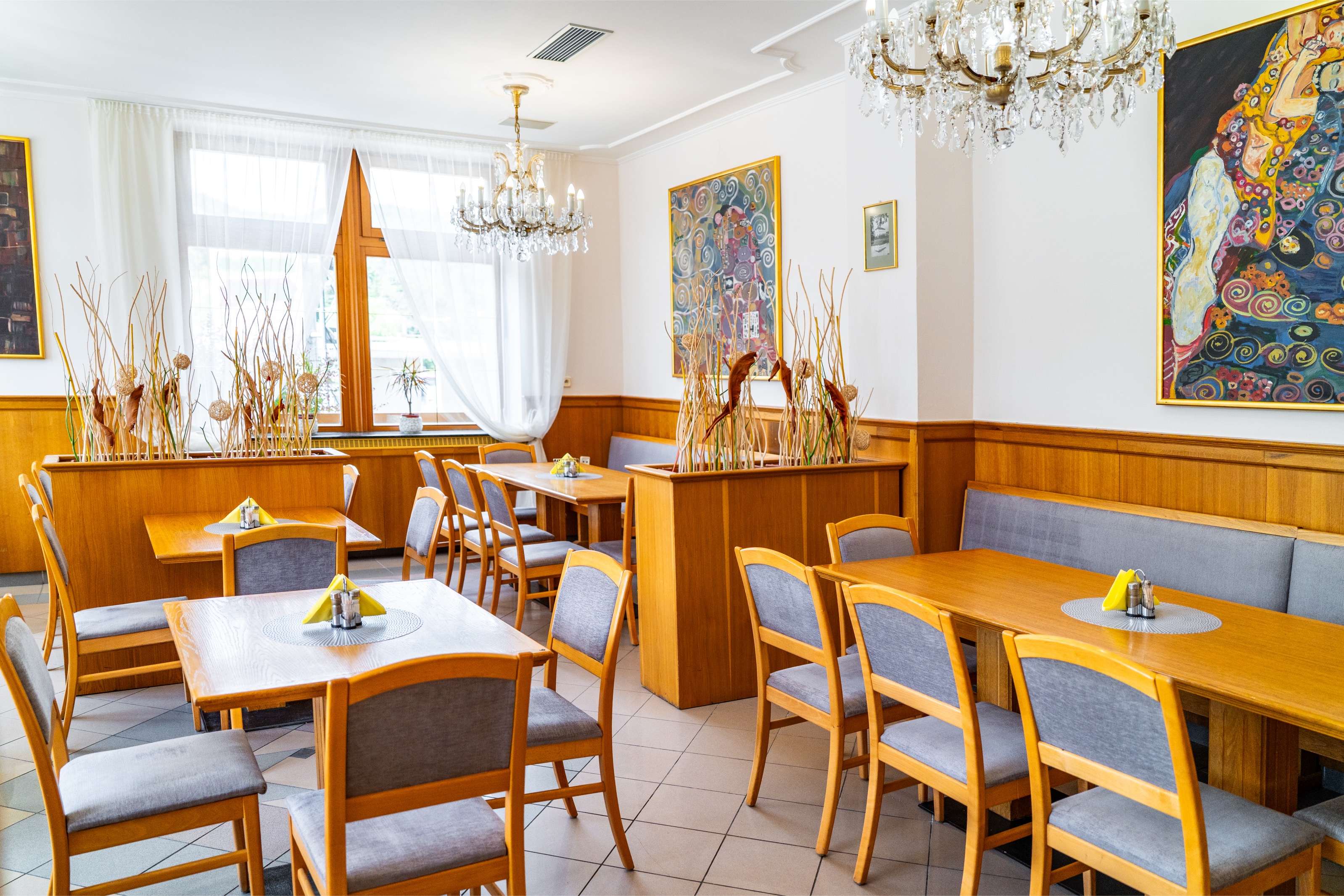 Bright restaurant dining area with wooden tables, gray cushioned chairs, decorative plants, crystal chandeliers, and colorful framed paintings on white walls.
