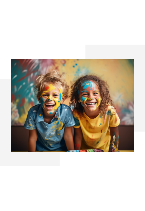 Two children smiling with colorful paint splattered on their faces and clothes against a bright background.
