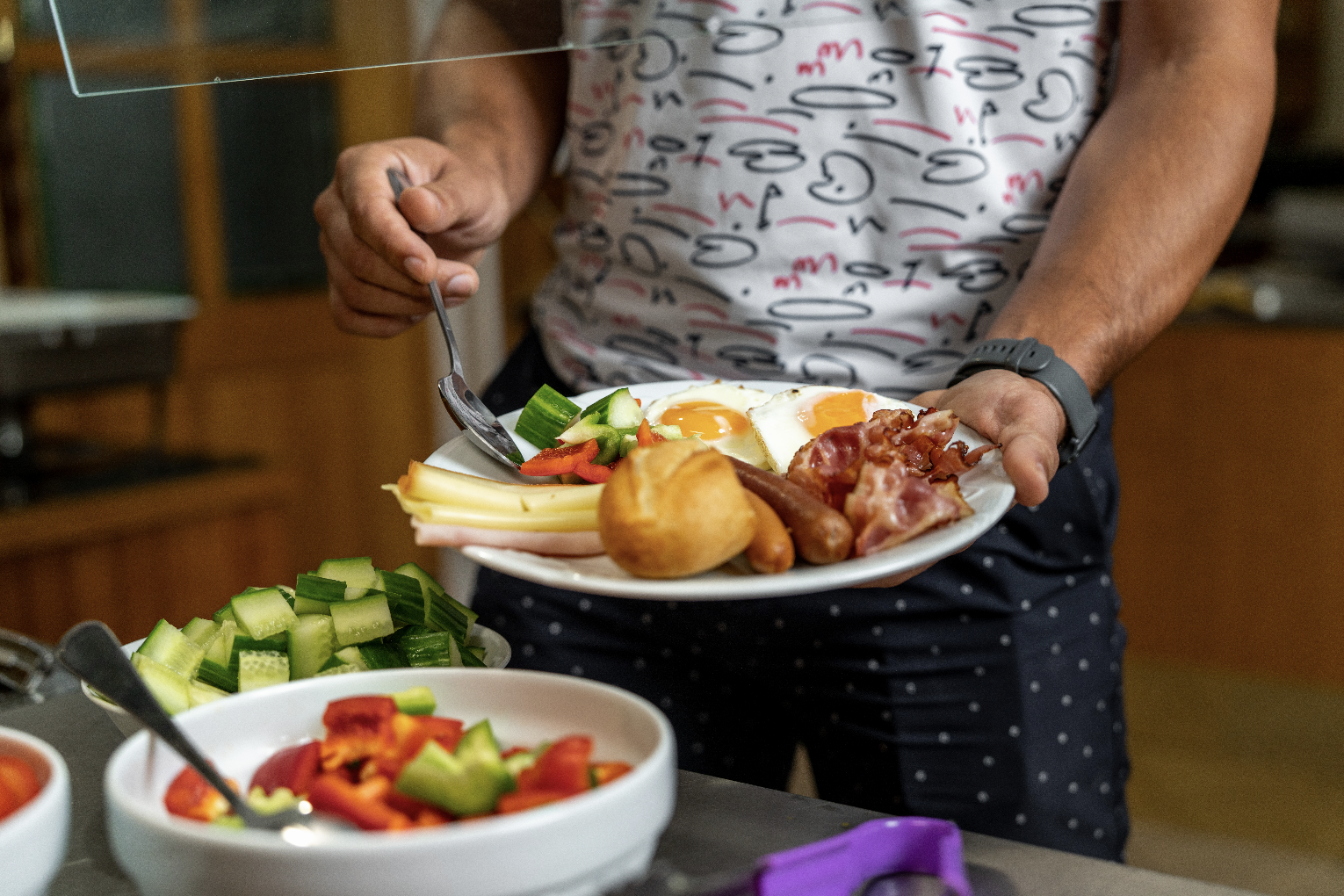 Person holding a plate with fried eggs, bacon, sausages, cheese, sliced cucumber, and a bread roll.