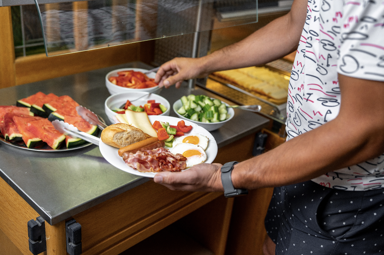Person serving a breakfast plate with fried eggs, bacon, sausage, cheese, salad, and bread at a buffet with watermelon slices and vegetable bowls.