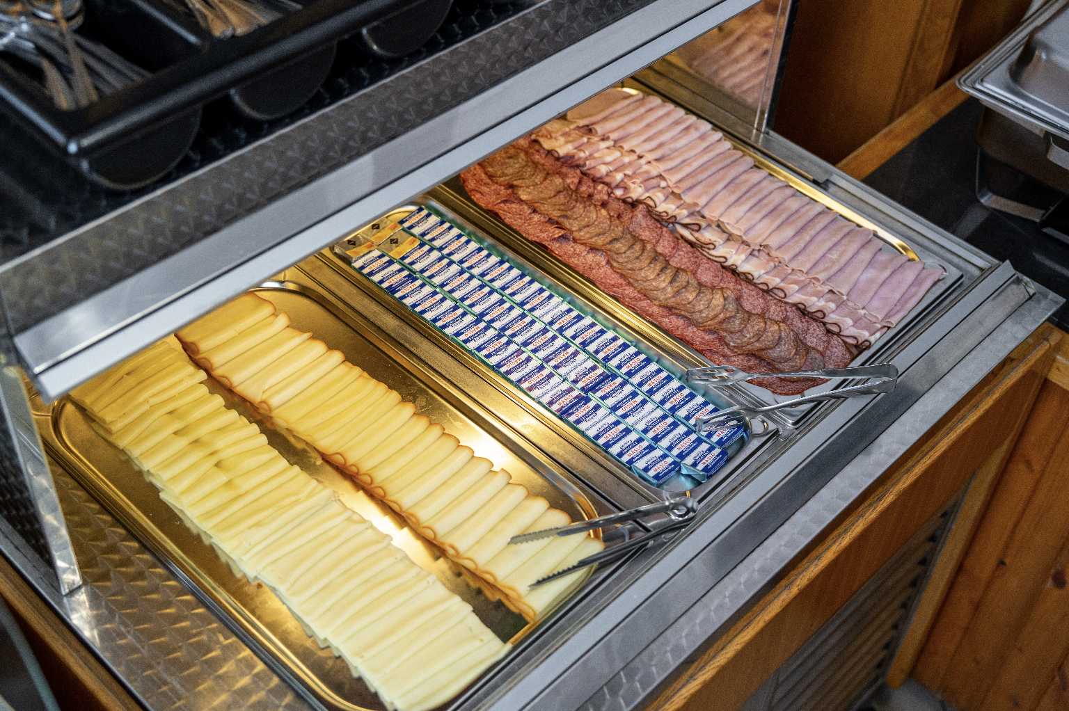 Buffet tray with neatly arranged sliced cheese, individually wrapped butter packets, and various sliced deli meats including ham and salami.