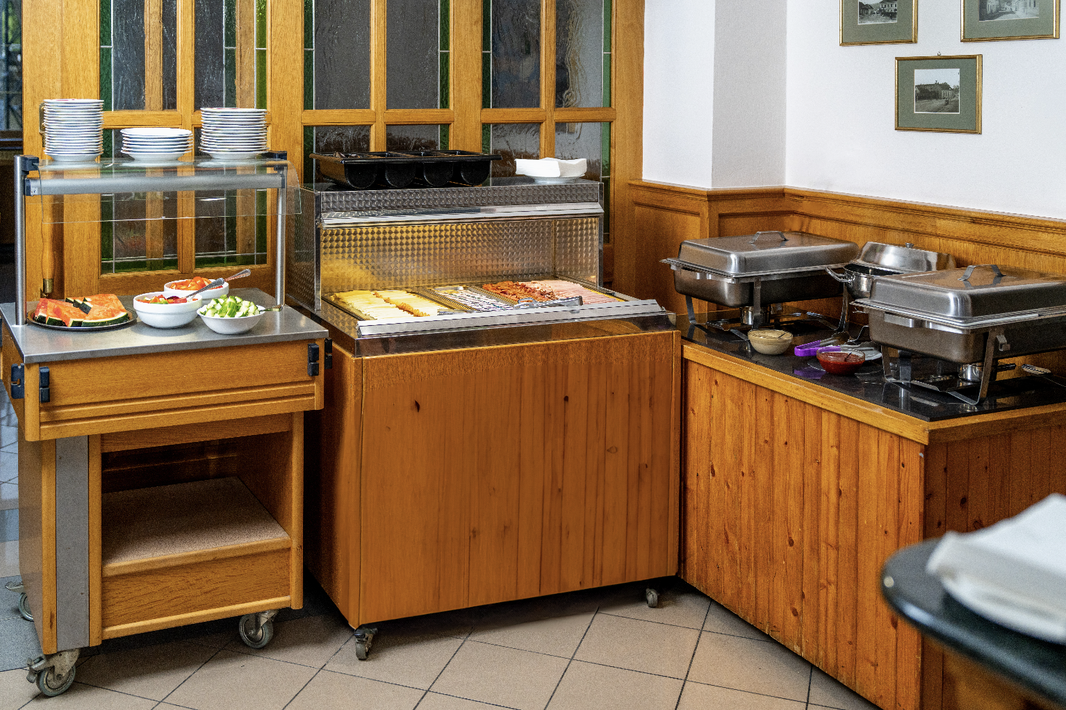 Buffet setup with plates, bowls of fresh fruit and salad, cheese and meat slices in a refrigerated display, and covered chafing dishes on a wooden counter.