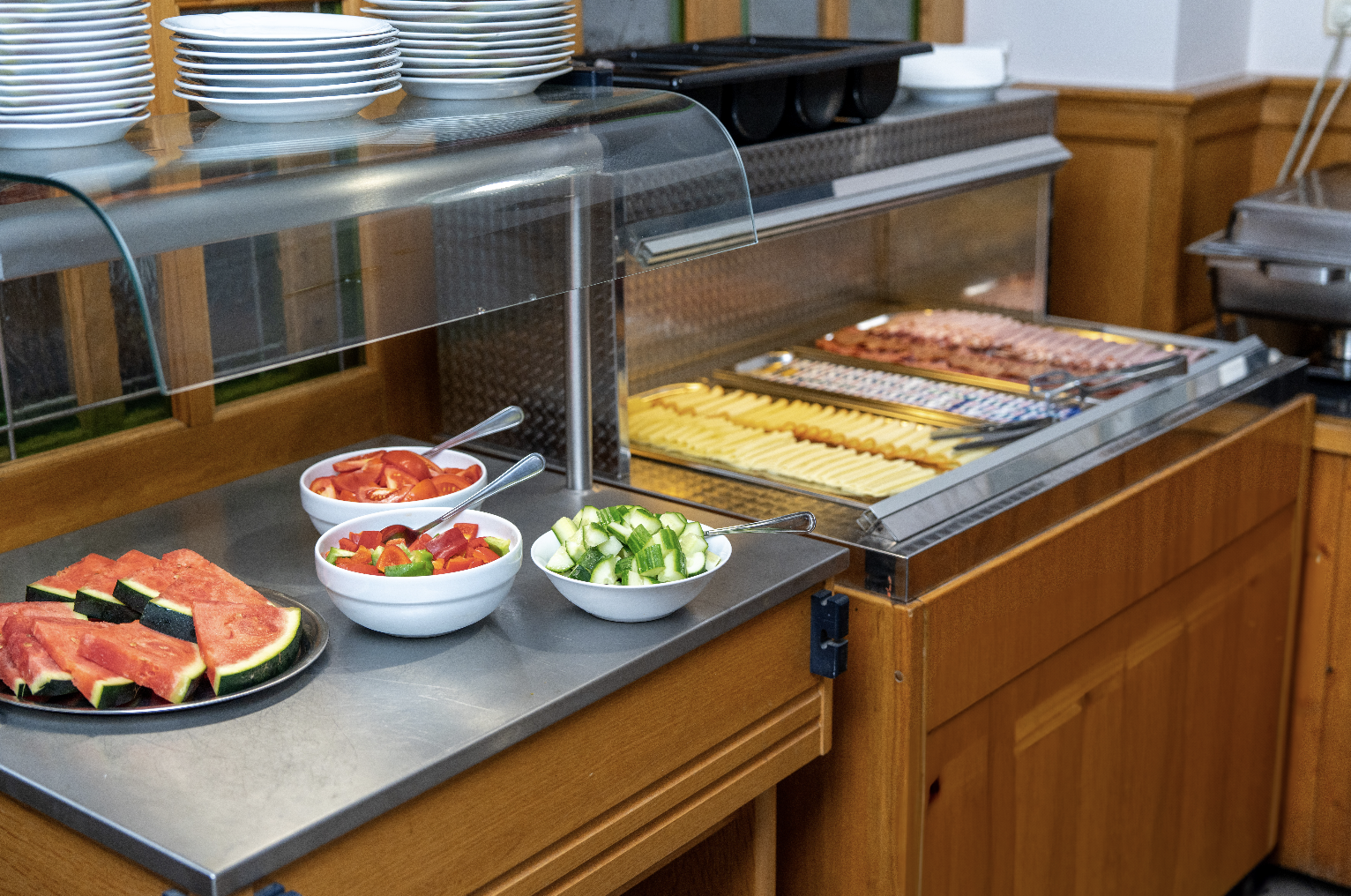 Buffet table with plates, bowls of chopped vegetables, slices of watermelon, and trays of sliced cheese and meats.